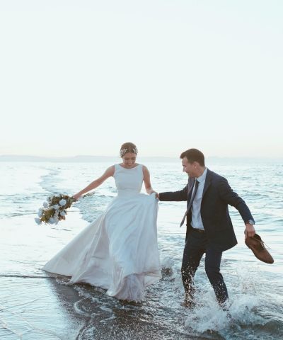 A bride in a white gown wades into the surf with a groom in a suit holding her bouquet, on a windy beach shore.