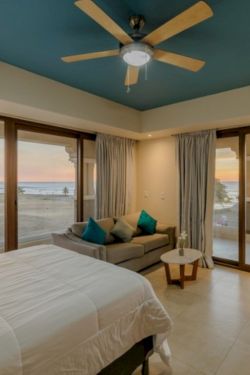 Coastal bedroom with sea view, sliding glass doors, cozy sofa and bed, and a wooden ceiling fan overhead.