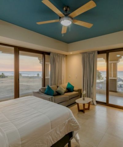 Coastal bedroom with sea view, sliding glass doors, cozy sofa and bed, and a wooden ceiling fan overhead.