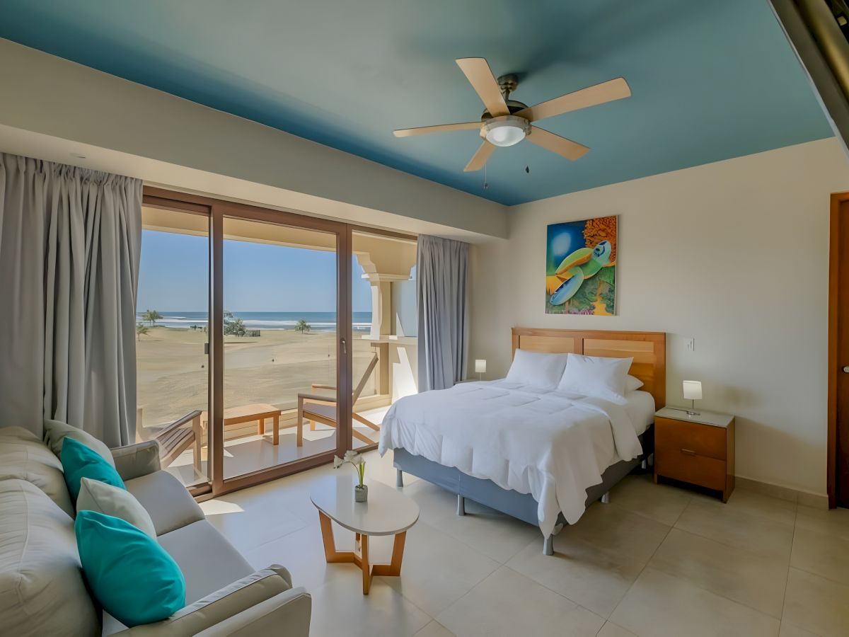 A beachside bedroom with a ceiling fan, large windows to the ocean, a bed, seating, and colorful wall art.