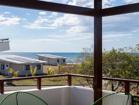 Balcony view framed by dark beams, overlooking a white villa courtyard, tropical trees, and the blue ocean under cloudy sky.