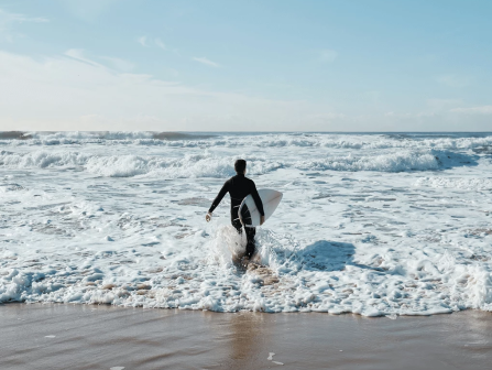 A surfer paddles into foamy waves on a sandy beach under a bright blue sky.