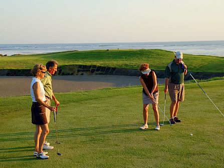 Four golfers line up their putts on a sunny seaside green with the ocean and waves in the background.