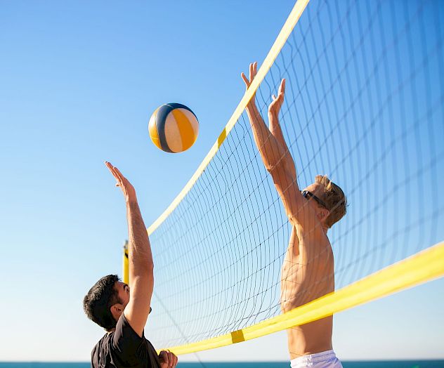 Two players jump at a beach volleyball net, one reaching to spike a yellow-and-blue ball against the other.