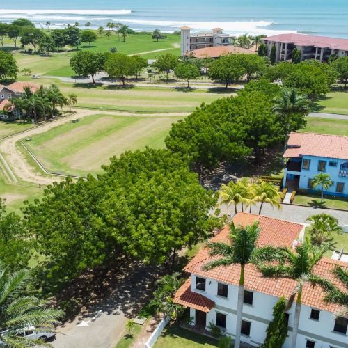 Aerial view of a seaside resort: red-roof buildings, lush green lawns and palm trees along a winding coastline.