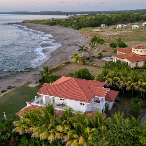 Aerial view of a tropical beachfront resort with red-roof villas, palm trees, and a quiet sandy shore along the ocean.