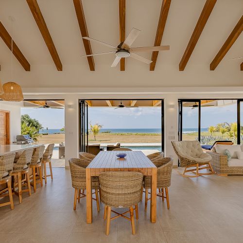 Beachside living room with vaulted wooden beams, ceiling fans, wicker dining & seating, and ocean view through sliding doors.