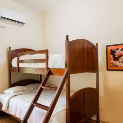 A bright bedroom with a wooden bunk bed, wall AC, open window, and a framed floral picture on the wall.