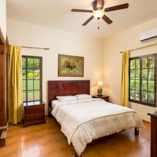 A sunny bedroom with a wooden bed, ceiling fan, yellow curtains, framed art, and large windows with greenery outside.