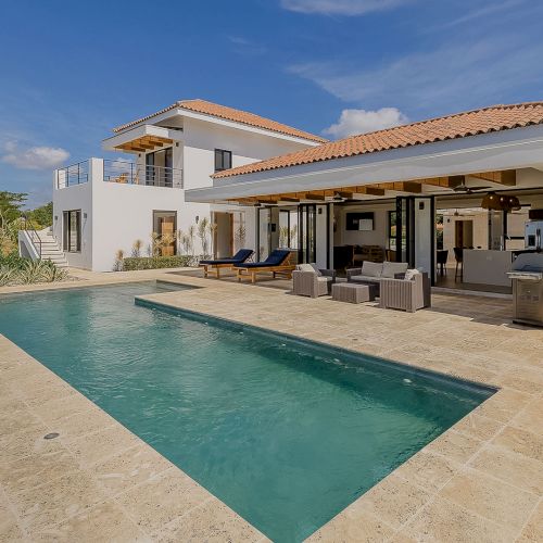 Modern white villa with a blue swimming pool, patio lounge furniture, and tiled outdoor kitchen under a blue sky.