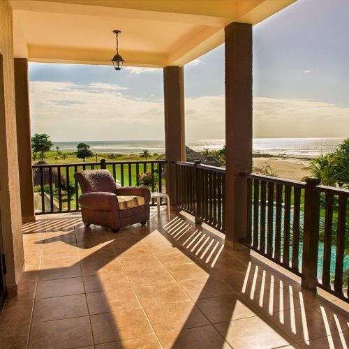 A sunny beachfront balcony with tiled floor, armchair, and railings overlooking the ocean and palms.