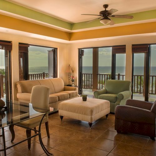 A seaside living room with couches, chairs, a glass table, and a ceiling fan, looking out at the ocean through sliding doors.