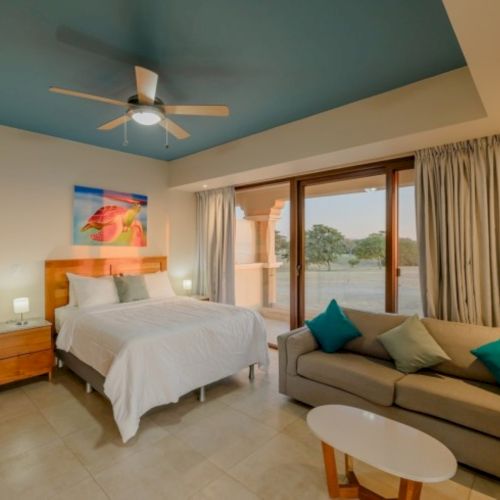 A bright bedroom with a ceiling fan, double bed, sofa, curtains, and large glass doors showing an outdoor view.