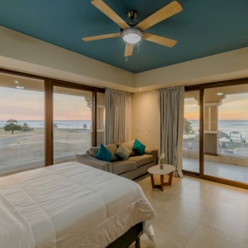 A seaside bedroom with a ceiling fan, plush bed, and lounge seating, framed by big windows showing the ocean and sunset.