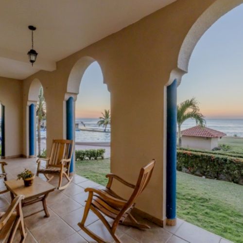 A seaside patio with arched columns, blue posts, wooden chairs, a lawn, and a sunset ocean view.
