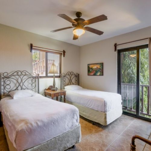 Bright bedroom with two beds, a ceiling fan light, and large windows with curtains showing greenery.