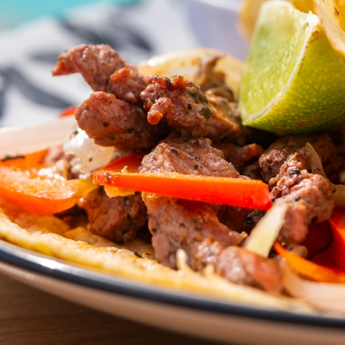 A sizzling plate of chopped beef with carrots and onions, tucked into a warm bread/taco shell, topped with herbs and a lime wedge.