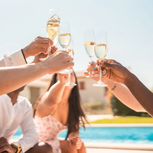 Friends clink champagne flutes outdoors by a pool, celebrating a sunny toast together with bubbles and smiles.