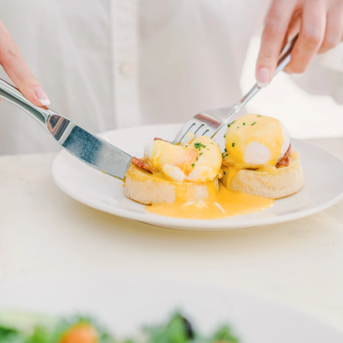 A person serves eggs Benedict with hollandaise on toast using a knife and fork at a breakfast table.