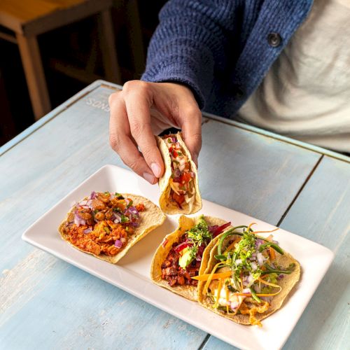 A hand holds a stuffed taco with toppings (meat, shredded veggies, salsa) over a plate of more tacos, ready to eat.