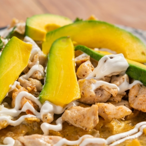 A plate of nachos/taco bites topped with shredded chicken, melted cheese, crema, and avocado and green pepper slices.