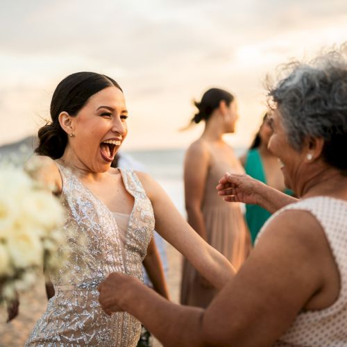 Friends celebrate at a sunny beach sunset, as a woman in a sparkly dress laughs and dances with others nearby.