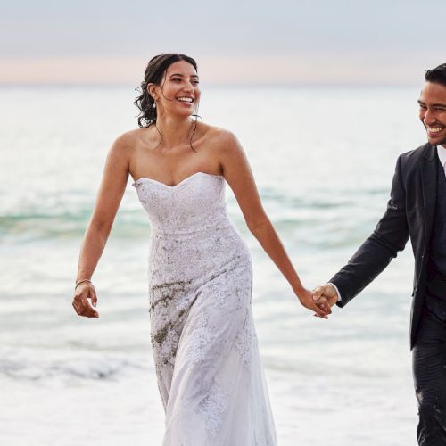 A smiling couple in wedding attire holds hands and walks along the beach at sunset, celebrating their special day.