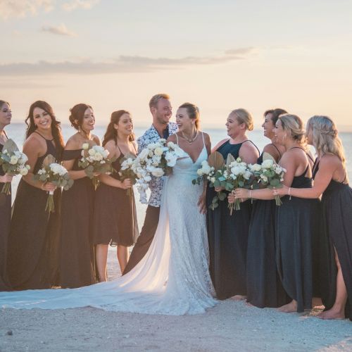 A bride and bridal party stand on a beach at sunset, smiling and holding bouquets.