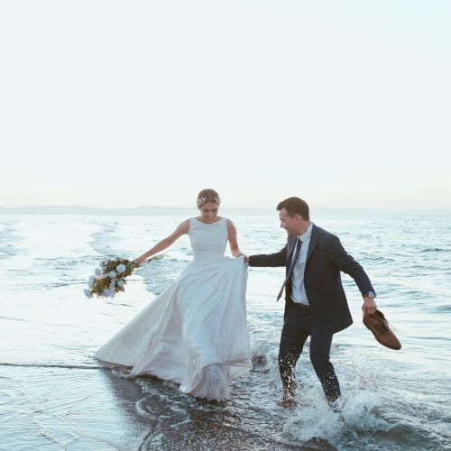 A bride and groom at the beach step into the waves with a bouquet, celebrating their wedding day.