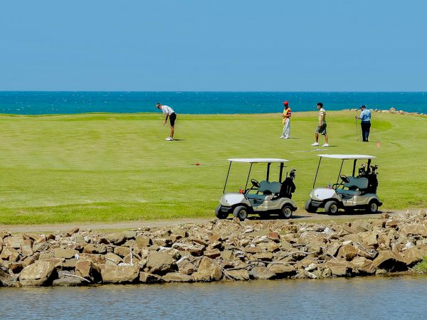 A group of golfers plays on a sunny seaside course, with two golf carts parked in the foreground and players lining up shots.