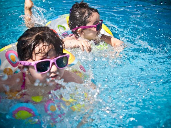 Two kids splash in a blue pool wearing swim goggles and floating toys, enjoying a sunny swim.