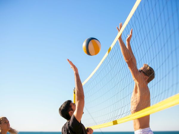 Beach volleyball: players reach over the net to spike a ball under a clear blue sky.
