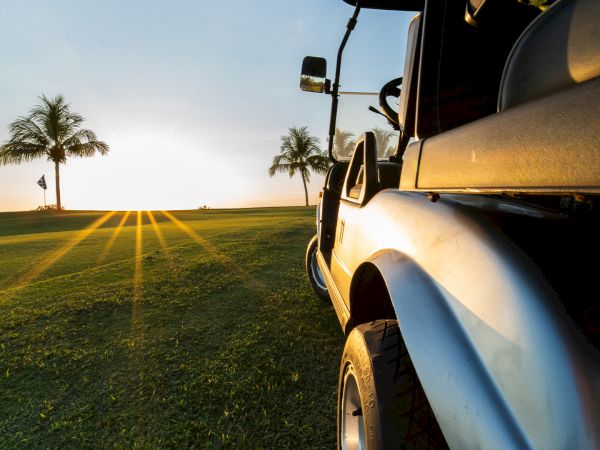 A white car is parked on a grassy field at sunset, with palm trees and long light rays in the distance.