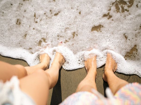 Bare feet play in foamy ocean waves on a sandy beach, with water washing over toes and ankles.