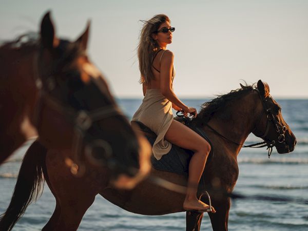 A woman in sunglasses rides a horse along the ocean at sunset, silhouetted by the waves and warm light.