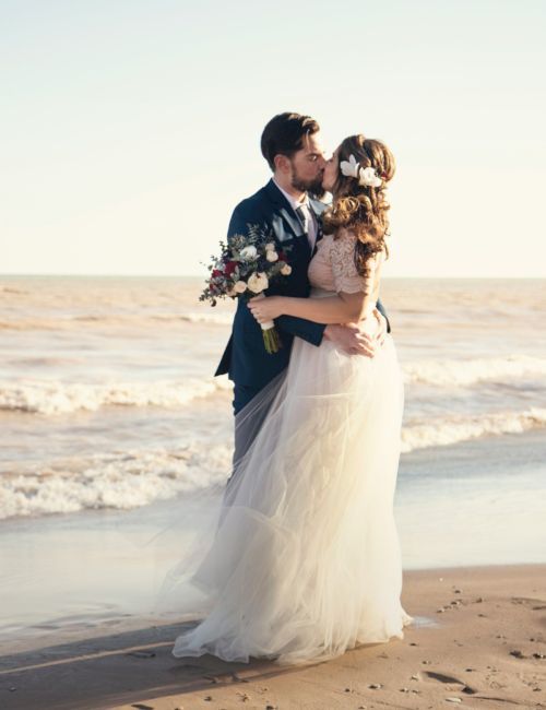 A bride and groom kiss on a sunny beach, holding a bouquet, with waves behind them during their wedding photo shoot.