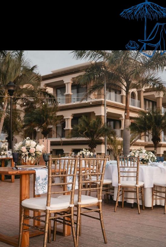 An outdoor wedding setup with gold chairs, floral centerpieces, and a tropical resort building with palm trees behind it.