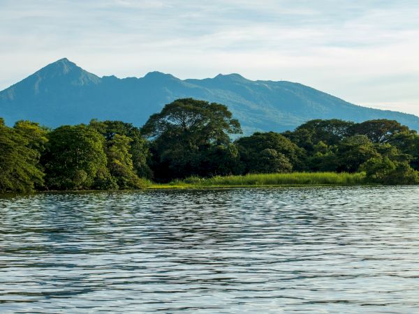 A tranquil lake in the foreground, lush green trees along the shore, and a blue mountain ridge under a pale sky.