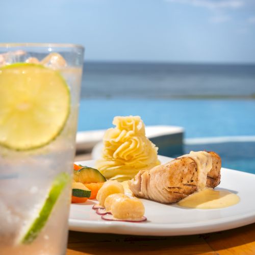 A sunny seaside meal: grilled chicken with creamy sauce, mashed potato, veggies, and a lemon drink beside the ocean view.