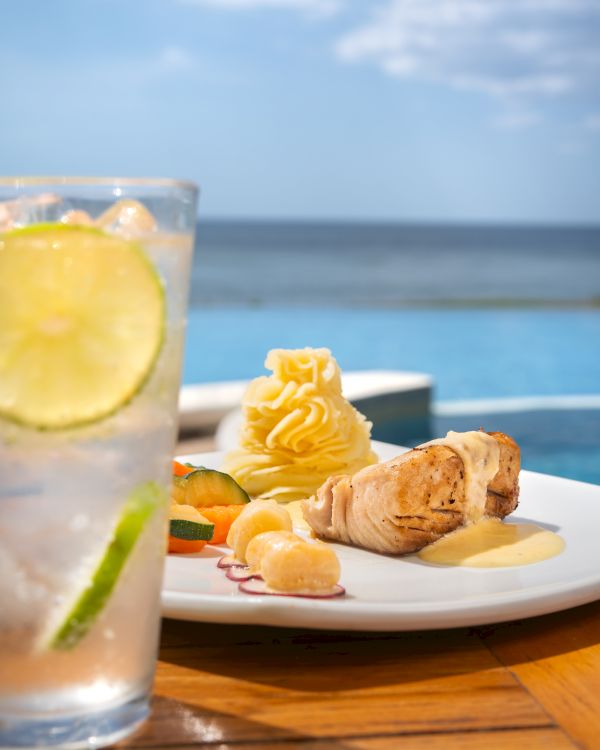 A sunny seaside meal: grilled chicken with creamy sauce, mashed potato, veggies, and a lemon drink beside the ocean view.