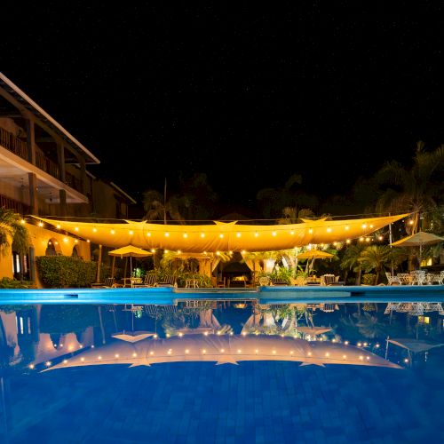 Nighttime hotel pool with warm string lights strung under a canopy, lounge chairs, palm trees, and reflections on the water.