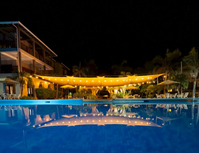 Nighttime hotel pool with warm string lights strung under a canopy, lounge chairs, palm trees, and reflections on the water.