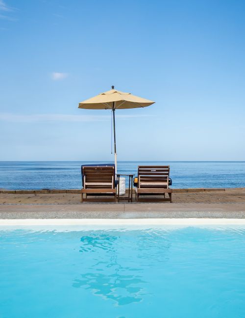 Two deck chairs under a beige umbrella by the ocean, beside a blue pool under a clear sky.