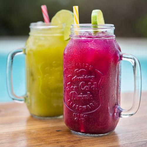 Two mason jars of bright yellow and purple smoothies with striped straws and citrus slices on a wooden table.