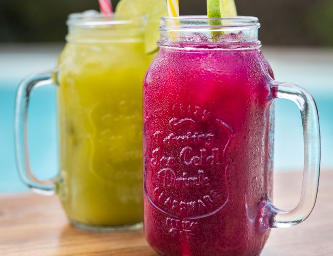 Two mason jars of bright yellow and purple smoothies with striped straws and citrus slices on a wooden table.
