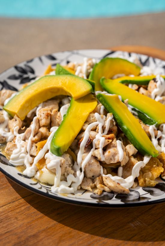A plate of loaded nachos with diced chicken, white drizzle, and fresh avocado slices, all on a toasted base.